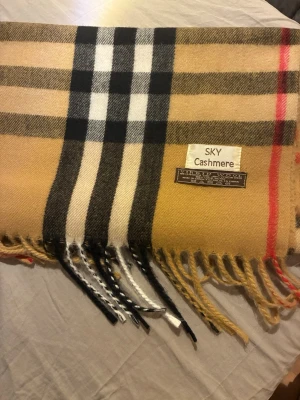 Beige rutig cashmereväst från SKY - Säljer en beige väst med klassiskt rutigt mönster i svart, vitt och rött från SKY Cashmere. Västen har fransar längs nederkanten och är tillverkad i mjuk cashmere och virgin wool. Perfekt för dig som gillar tidlös och stilren design.