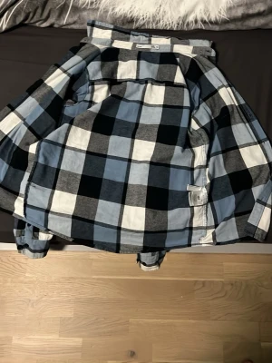 Rutig overshirt i blå och svart - Säljer en snygg overshirt från lager 157 med stort rutigt mönster i blått, svart, grått och vitt. Jackan har klassisk krage, knappar framtill och på ärmsluten samt en bröstficka med lock. Perfekt att slänga över en shorta  för en chill look. 