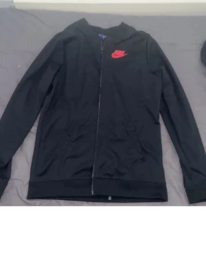 Svart bomberjacka från Nike - Svart bomberjacka från Nike med röd logga på bröstet. Jackan har dragkedja framtill och långa ärmar. Perfekt för dig som gillar sportig och clean stil. Materialet känns mjukt och lätt, och jackan har en klassisk passform.