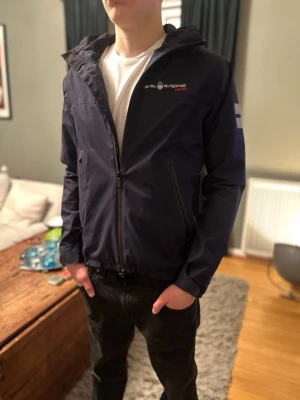Mörkblå Sail Racing vindjacka - Snygg mörkblå vindjacka från Sail Racing med huva och dragkedja framtill. Jackan har logga på bröstet och reflekterande detaljer på ärmen och ryggen. Två snedställda fickor med dragkedja och en clean, sportig look.