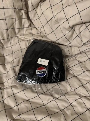Svart Pepsi mössa med broderad logga - Svart ribbstickad mössa med broderad Pepsi-logga framtill i rött, vitt och blått. Klassisk form med uppvikt kant, perfekt för dig som vill ha en enkel men snygg accessoar. Materialet känns mjukt och värmande. Den är aldrig använd 