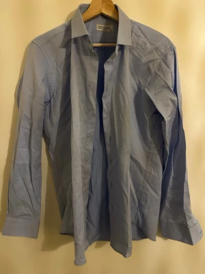 Blå skjorta från Suvari med knappar - Snygg blå skjorta från Suvari med klassisk krage och långa ärmar. Skjortan har diskret mönster och knäppning framtill med små knappar. Perfekt för dig som gillar stilrena och enkla plagg. Materialet känns lätt och bekvämt mot huden.