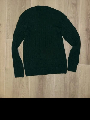 Mörkgrön stickad tröja med rund hals - Säljer en mörkgrön stickad tröja med rund hals och ribbade muddar. Tröjan har ett klassiskt kabelstickat mönster och långa ärmar, perfekt för kyliga dagar. Enkel och stilren design som funkar till många olika outfits.