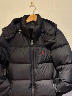 Svart pufferjacka från Polo Ralph Lauren - Svart pufferjacka från Polo Ralph Lauren med klassisk röd logga på bröstet. Jackan har hög krage, huva och flera dragkedjor framtill. Materialet är polyester och jackan har en quiltad design som ger en schysst volym. Perfekt för kalla dagar.