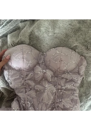 Ljuslila blommig bustierklänning Eilly Bazar - Supersöt kort klänning från Eilly Bazar i ljuslila med stora blommiga detaljer och paljetter. Axelbandslös bustiermodell med markerade sömmar och dragkedja i sidan. Perfekt för dig som vill sticka ut lite extra!