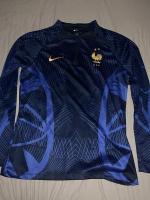 Inga skor på bilderna - Bilderna visar ett träningsset från Nike och franska landslaget, men inga skor finns med. Setet består av en långärmad tröja och byxor i marinblått med guldiga detaljer och mönster.