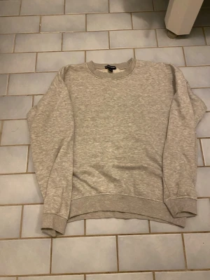 Grå sweatshirt från PrettyLittleThing - En basic grå sweatshirt från PrettyLittleThing i mjuk bomullsblandning. Tröjan har rund halsringning, ribbade muddar vid ärmslut och nederkant samt lång ärm. Perfekt för chill dagar eller när du vill ha en enkel och clean look.