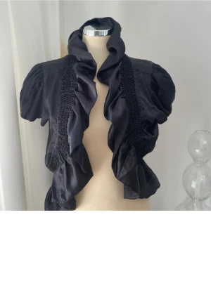 Svart volangblus bolero från Sisters Point - Supersnygg svart blus /bolero från Sisters Point med stora volanger framtill och puffiga korta ärmar. Materialet är glansigt och känns lyxigt, perfekt för att lyfta vilken outfit som helst. Smockdetaljer längs sidorna ger en cool och unik look.