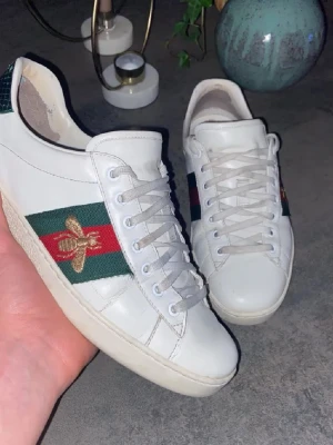 Gucci ace skor säg ditt pris - Säljer ett par vita Gucci sneakers i skinn med klassiska gröna och röda ränder på sidan, broderad gul bi och metallicröd samt metallicgrön häl med Gucci-logga. Skorna har snörning och platt sula, rund tå och cool streetstil. Perfekt för dig som gillar lyxiga detaljer och vill sticka ut.
