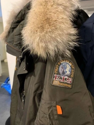Parajumpers parkas XL - Säljer en mörkgrön Parajumpers parkas med stor fluffig päls runt huvan och flera praktiska fickor. Jackan har patch på ärmen, dragkedja och knappar framtill samt orangea detaljer. Perfekt för kalla dagar och riktigt snygg streetstil.