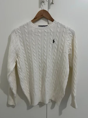 Vit kabelstickad tröja Polo Ralph Lauren - Vit kabelstickad tröja från Polo Ralph Lauren med klassisk rund hals och broderad logga på bröstet. Tröjan har långa ärmar och ribbade muddar vid ärmslut och nederkant. Perfekt för dig som gillar stilrena och tidlösa plagg.