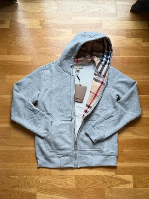 Burberry zip up grå S - En grå zip-hoodie från Burberry i mjukt bomullsmaterial med dragkedja framtill. Har en klassisk huva med det ikoniska rutmönstret på insidan och en broderad logotyp i form av en riddare på bröstet. Perfekt som vardagsplagg eller för en avslappnad men stilren look. Inga synliga defekter, i fint skick.