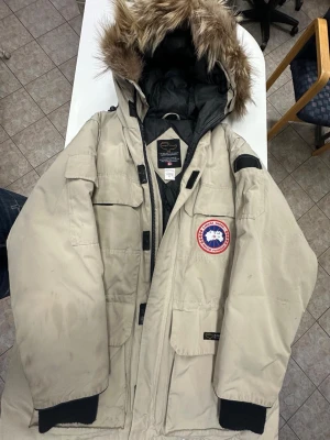 Beige dunjacka med päls från Canada Goose - Säljer en beige dunjacka från Canada Goose med svart quiltad insida och stor huva med päls. Jackan har flera fickor, dragkedja och klassisk patch på bröstet. Perfekt för kalla vinterdagar och har en riktigt snygg look med pälsdetaljen på huvan.