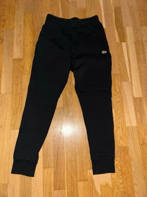 Svarta mjukisbyxor från Lacoste - Säljer ett par svarta mjukisbyxor från Lacoste med klassisk logga på låret. Byxorna har resår i midjan och muddar vid bensluten. Aldrig använda
