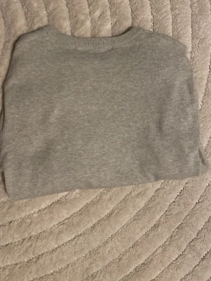 Ljusgrå stickad tröja - En enkel och stilren ljusgrå stickad tröja med rund halsringning och långa ärmar. Tröjan har ribbade muddar vid hals, ärmslut och nederkant. Perfekt basplagg för lager på lager under kyligare dagar. Den är lite liten i storlken 