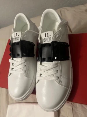 Valentino Rockstud Untitled sneakers - Säljer ett par vita Valentino Rockstud Untitled sneakers med svart band och ikoniska pyramidnitar över vristen. Skorna har snörning, rund tå och platt sula. Tillverkade i skinn med coola detaljer som ger en edgy vibe. Perfekt för dig som gillar lyxiga och unika sneakers.