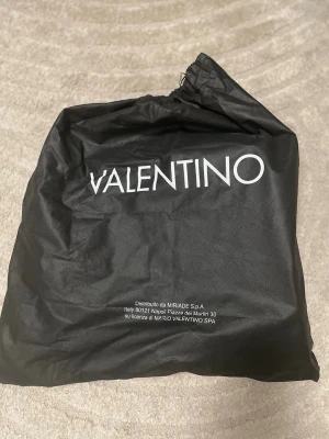 Svart ryggsäck från Valentino - Säljer en snygg svart ryggsäck från Valentino med diskret logomönster över hela väskan. Ryggsäcken har justerbara axelremmar med Valentino-tryck, ett stort huvudfack med innerficka och en praktisk ytterficka med dragkedja. Materialet är skinnliknande syntet och detaljerna är i svart. Skicka gärna frågor och bud!