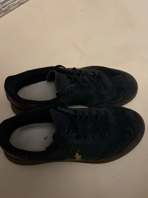 Mörkblå Polo Ralph Lauren sneakers - Snygga mörkblå sneakers från Polo Ralph Lauren med gröna detaljer på sidorna och guldlogga på plösen. Skorna har bruna gummisulor, snörning och bordeauxröd häl med Polo-tryck. Ovandelen är i mocka och insidan är mjukt fodrad för extra komfort.