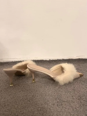 Beige tofflor med päls och spetsig tå - Säljer ett par beige tofflor med mjuk pälsdetalj över foten och spetsig tå. Skorna har låg klack med gulddetalj längst ner och öppen häl. Perfekt för dig som gillar unika och trendiga tofflor med lyxig känsla.