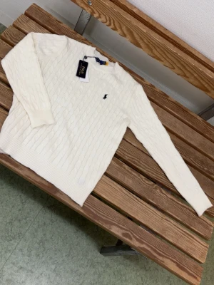 Vit kabelstickad tröja Polo Ralph Lauren - Vit kabelstickad tröja från Polo Ralph Lauren i storlek S. Tröjan har lång ärm, rund hals och den klassiska broderade loggan på bröstet. Mjuk och skön känsla med ribbade muddar vid ärmslut och nederkant. Perfekt för dig som gillar stilren och tidlös design.