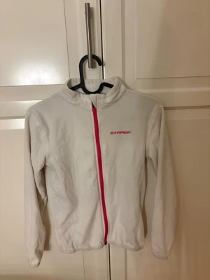 Vit fleecejacka från Everest - Säljer en vit fleecejacka från Everest med rosa dragkedja. Jackan har lång ärm, hög krage och är gjord i mjukt fleecematerial som värmer bra. Köp ju jag postar snabbt!💕💗