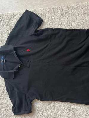 Svart piké från Polo Ralph Lauren - Klassisk svart pikétröja från Polo Ralph Lauren i bomull med röd broderad logga på bröstet. Modellen har krage, korta ärmar och två knappar vid halsen. Custom fit för en snygg passform. Perfekt till jeans eller shorts.