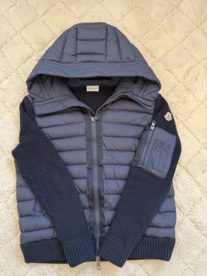 Svart  pufferjacka från Moncler - Snygg svart pufferjacka från Moncler med stickade ärmar och huva. Jackan har quiltad framsida, tvåvägsdragkedja och en ficka med dragkedja på ärmen. Materialmix av polyester och stickad ull ger en cool look. Perfekt för dig som gillar streetstyle.