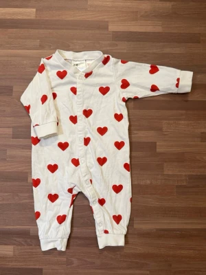 Lång body med röda hjärtan - Supergullig vit overall från H&M med röda hjärtan i storlek 62 (2-4mån). Långa ärmar och ben, samt praktiska tryckknappar framtill för enkel påklädning. Endast upptvättat och är i nyskick.