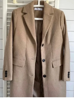 Beige kappa från Ellos med knappar - Snygg beige kappa från Ellos med klassisk krage och svarta knappar framtill. Jackan har långa ärmar, fickor med lock och är tillverkad i ett ullblandat material som ger en stilren look. Perfekt för dig som gillar enkel och tidlös design.