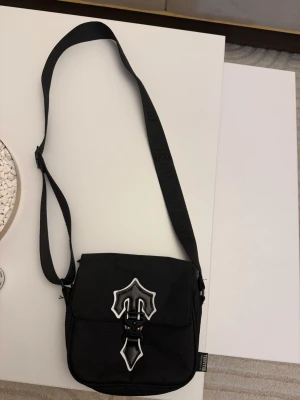 Svart axelväska från Trapstar - Säljer en svart axelväska från Trapstar med justerbar rem och coolt emblem i svart och vitt på framsidan. Väskan har ett spänne framtill och är tillverkad i slitstark syntet. Perfekt för att bära dina viktigaste grejer med stil.