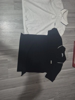 Svart och vit polotopp från BOSS - Två stilrena polotoppar från BOSS, en i svart och en i vit med kontrasterande tunna ränder på krage och ärmslut. Klassisk design med korta ärmar, knappar vid halsen och broderad BOSS-logga på bröstet. Perfekt för en clean och snygg look. I den sista bulden ser ni att den vita polon är lite smutsig men drt går säkert bort med tvätten, ej säker på det.