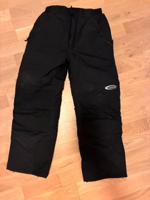 Svarta täckbyxor från Cover - Svarta täckbyxor från Cover med elastisk midja och kardborreknäppning. Utrustade med dragkedjor vid fickorna och ett värmande foder. Perfekta för kalla dagar och vinteraktiviteter. Snyggt broderad logga på benet.