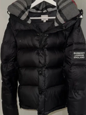 Svart pufferjacka från Burberry - Svart pufferjacka från Burberry med klassiskt rutigt foder i grått, svart och rött. Jackan har dragkedja och knappar framtill, samt en patch med Burberry London England på ärmen. Perfekt för kalla dagar och har en riktigt snygg street vibe.