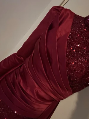 Vinröd glittrig långklänning Giffniseti - Vinröd långklänning från Giffniseti med glittriga paljetter över hela klänningen och ett elegant omlott-draperat tyg i satin. Klänningen har långärmade ärmar och en figurnära passform som ger en lyxig känsla. Perfekt för bal eller festliga tillfällen.