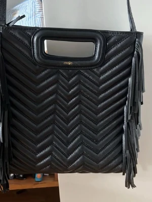 Svart maje väska  - Säljer en svart axelväska från Maje i quiltat skinn med snyggt zigzag-mönster. Väskan har fransar längs sidorna och ett stilrent handtag upptill. Perfekt för dig som gillar detaljer och vill ha en unik väska med cool vibe. Inga defekter och inga tecken på användning. Försiktigt använd också💖