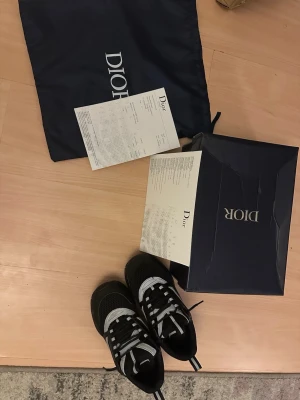 Svarta sneakers från Dior - Säljer ett par svarta Dior sneakers med vita detaljer och snörning. Skorna har en rund tå och platt sula, tillverkade i mesh och syntetmaterial. Medföljer originalkartong och dustbag med Dior-logga. Perfekt för dig som gillar lyxiga och stilrena sneakers.