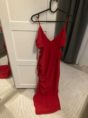Röd långklänning från ZARA - Säljer en röd långklänning från ZARA med smala axelband och v-ringning. Klänningen har en snygg drapering på sidan och stängs med dragkedja i ryggen. Materialet är mjukt och följsamt, perfekt för en statement-look.