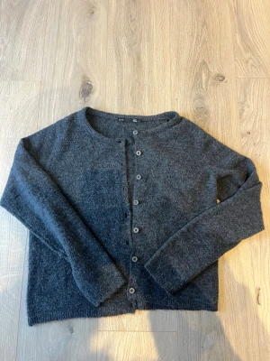 Mörkgrå stickad kofta från 157 strl S - Mysig mörkgrå stickad kofta från 157 i storlek S. Koftan har rund halsringning och knäppning med grå knappar framtill. Perfekt att slänga på sig när det är lite kyligt ute. Enkel och stilren design som funkar till många olika outfits.