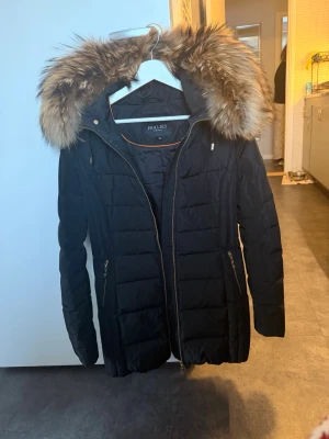 Svart pufferjacka med päls från Hollies - Snygg svart pufferjacka från Hollies med fluffig päls på huvan och guldfärgade dragkedjor. Jackan har quiltad design, lång ärm och praktiska fickor med dragkedja. Perfekt för kalla dagar och har en stilren badge på ärmen.Knappt använd,så nyskick och toppenfin.Dyr i inköp