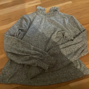 Grå långärmad tröja från Vero Moda - Mysig grå långärmad tröja från Vero Moda med volangkant vid halsen och knapp i nacken. Tröjan har ribbade muddar vid ärmslut och är tillverkad i mjukt stickat material. Perfekt för kyliga dagar och enkel att matcha med jeans eller kjol.