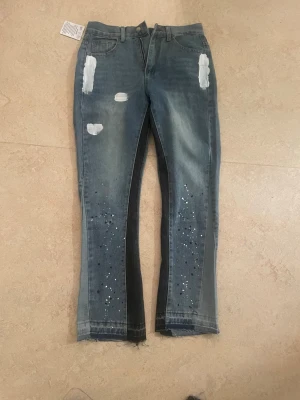 Gallery Dept. jeans med färgstänk i blå - Unika jeans från Gallery Dept. i blå och svart denim med handmålade vita färgstänk och slitna detaljer. Modellen har raka ben med croppad och rå kant nedtill, samt klassisk femficksdesign. Märkets patch syns både invändigt och på etiketten.