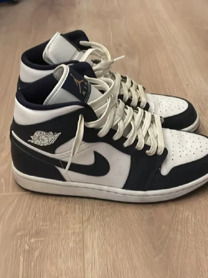Nike Jordan 1 Highs i marinblå och vit - Säljer ett par Nike Jordan 1 Highs sneakers i marinblått och vitt med klassisk snörning och rund tå. Skorna har den ikoniska Jordan-loggan på sidan och tungan, samt en platt sula. Perfekt för dig som gillar streetwear och vill ha en snygg och tidlös look.