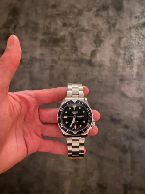 Helt Ny Seiko Mod Sports 5 - Säljer denna helt nya Seiko Mod Sports 5 klockan med svart urtavla som visar både datum samt veckodag, och som har en roterande bezel i svart färg✅  Klockan har en batteridriven Quartz movement, och den kommer med en klocklåda✅  Vid fler frågor eller funderingar skicka gärna ett pm, Mvh