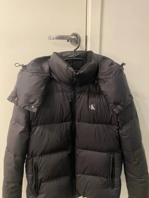 Svart pufferjacka från Calvin Klein Jeans - Svart pufferjacka från Calvin Klein Jeans med huva och dragkedja framtill. Jackan har CK-logga på bröstet och två fickor med dragkedja. Den är vadderad och har en clean, modern look som passar perfekt när det är kallt ute.