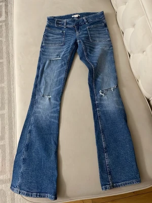 Blå bootcut jeans med slitningar - Snygga blå jeans med bootcut-snitt och slitna detaljer på båda benen. Jeansen har stora framfickor och klassiska bakfickor. Materialet är jeans och färgen är en tvättad blå ton med ljusare partier. Perfekt för dig som gillar en avslappnad och trendig stil.