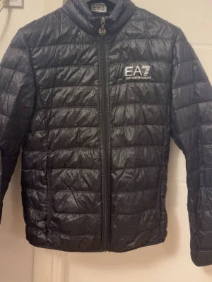 Svart quiltad pufferjacka EA7 - Svart pufferjacka från EA7 Emporio Armani med quiltad design och dragkedja framtill. Jackan har hög krage, två sidofickor och EA7-logga på bröstet. Materialet är glansigt och känns lätt, perfekt för kyliga dagar. Den var använde i cirka 2 månader förra året, har legat upphängd och orörd sedan dess.