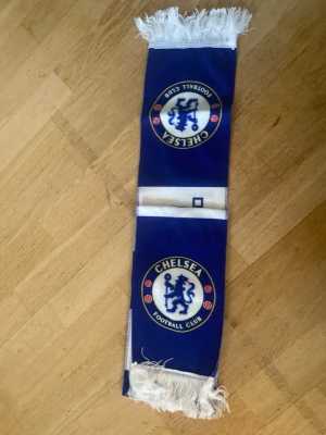 Chelsea F.C. halsduk i blått och vitt - Chelsea F.C. supporterhalsduk i blått och vitt med klubbens logga på båda ändarna och texten CHELSEA F.C. i mitten. Halsduken har vita fransar på kanterna och är tillverkad i mjukt syntetmaterial. Perfekt för att visa ditt stöd på matchen eller hemma.