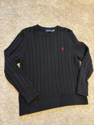 Svart kabelstickad tröja Polo Ralph Lauren - Klassisk svart kabelstickad tröja från Polo Ralph Lauren med rund hals och röd broderad logga på bröstet. Tröjan har långa ärmar och ribbade muddar vid ärmslut och nederkant. Perfekt för lager på lager under höst och vinter.          Vid frågor är det bara att höra av sig! 