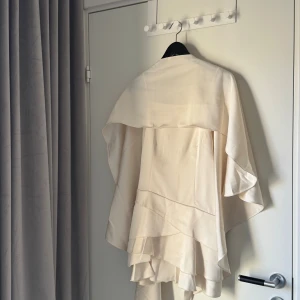 Champagnefärgad omlottklänning med volanger - Säljer en helt ny aldrig använd champagnefärgad kortklänning med tunna axelband och snygga volangdetaljer i flera lager. Materialet är mjukt och har en lätt glans. Kan även hämtas upp i Vasastan 