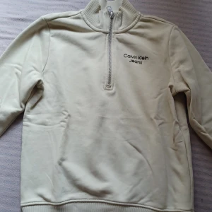 Beige Calvin Klein half zip sweatshirt - Snygg beige - grön  sweatshirt från Calvin Klein Jeans med halv dragkedja och broderad logga på bröstet. Tröjan har ribbade muddar och hög krage, perfekt för lager-på-lager. Tillverkad i mjuk bomullsmix och har en clean, stilren look. För barn str 140 - 10 år